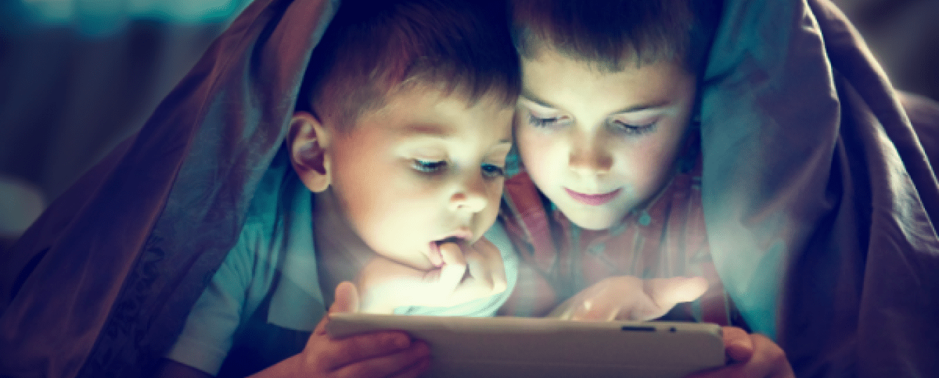 Twee kinderen liggen onder een deken en kijken zijn samen op een ipad