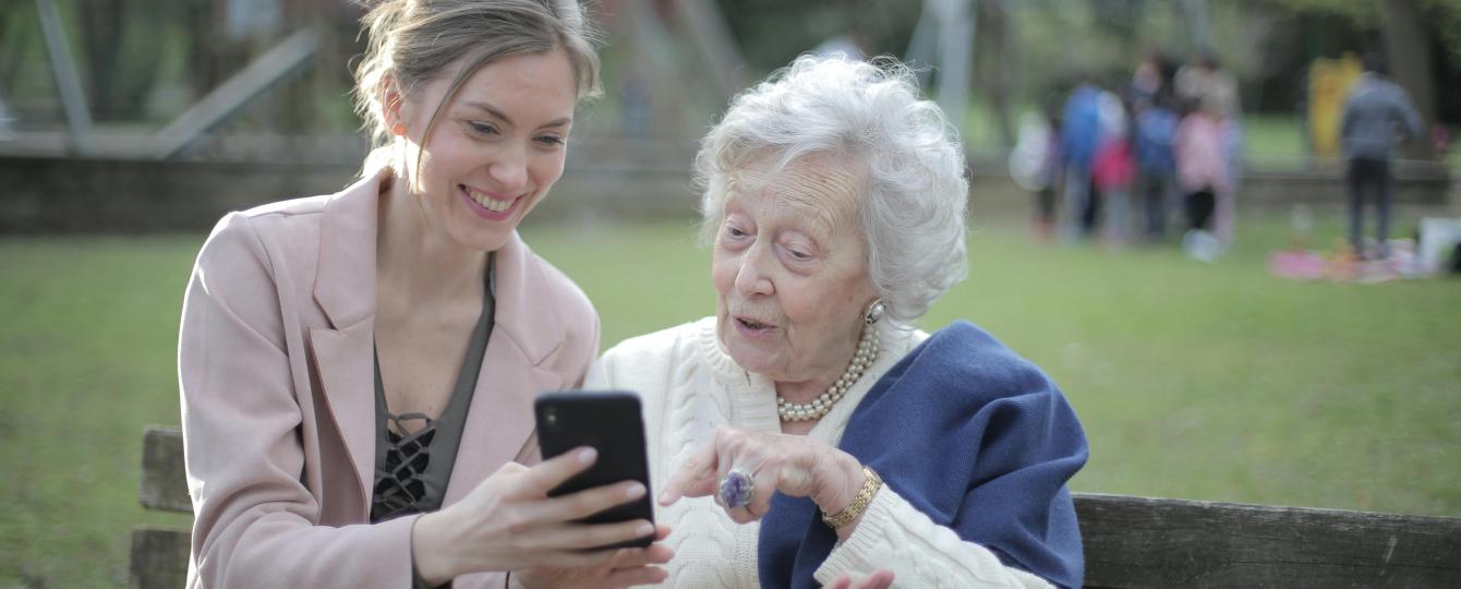 Jongere vrouw en ouderen vrouw zitten buiten op een bank en kijken samen naar een smartphone.