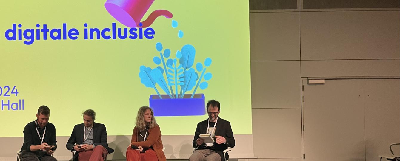 Groot scherm met presentatie met daarvoor vier mensen die op een stoel zitten